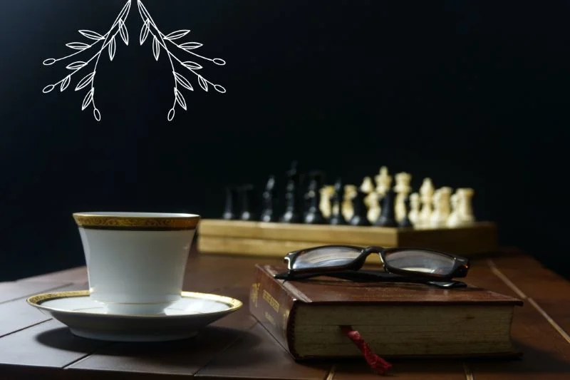A table with a cup, glasses on top of a book and a chessboard in the background.
