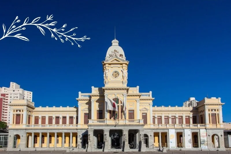 Fachada do museu, um grande edifício eclético amarelo e branco, com uma torre central. Céu azul ao fundo.