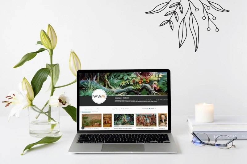 Computador sobre uma mesa mostrando o website da coleção on-line sobre estampas culturais. Na mesa, um vaso de flores, um livro, uma vela e óculos.